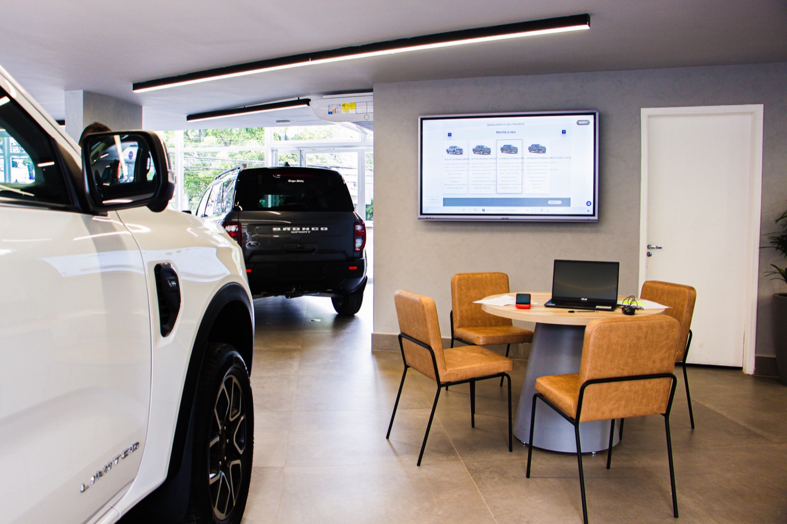 Monitor na parede da área de atendimento da concessionária Ford, com mesa e cadeiras em primeiro plano