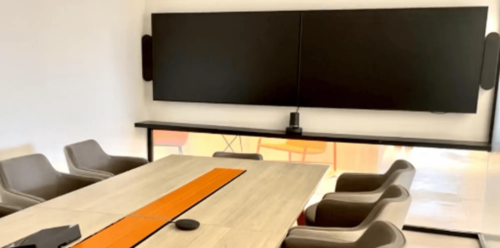 Dois monitores profissionais lado a lado em uma mesa de reunião, com um painel de LED ao fundo